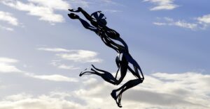 Artistic metal sculpture of a jumping figure set against a cloudy sky in daylight.