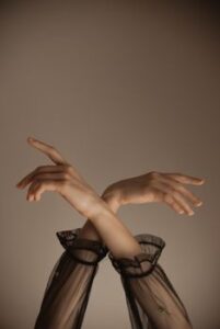 Crop anonymous female dancer in transparent black dress keeping hands up while performing sensual dance movement against gray background
