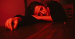 A pensive woman rests her head on a table, bathed in red lighting, reflecting emotion.